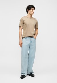 Homme portant une chemise beige à manches courtes, jean large bleu clair, chaussures noires, debout avec une main dans la poche, regardant à droite.