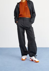 Svarta cargo-byxor med fickor, tillsammans med en orange strukturerad stickad tröja och en svart jacka. Skorna är vita med orange detaljer.