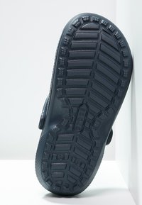 Semelle de chaussure en caoutchouc noir avec un motif texturé pour l'adhérence. Présente des sections surélevées et un logo circulaire au centre.