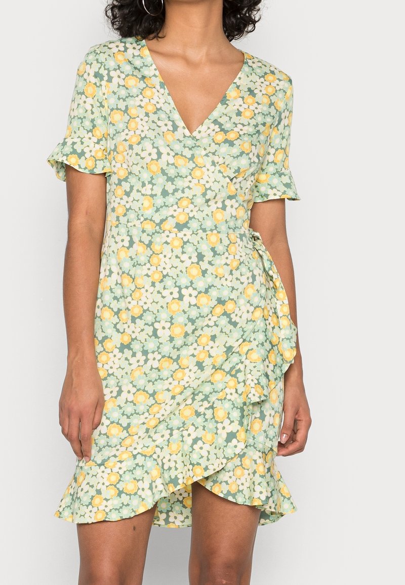Femme portant une robe portefeuille verte à manches courtes avec imprimé floral jaune et blanc et ourlet volanté sur un fond uni.