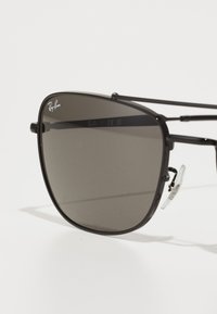 Ochelari de soare de tip black metal cu lentile întunecate. Cadru pătrat, dotat cu tampoane de nas din silicon și cu logo-ul Ray-Ban pe lentila din stânga sus.