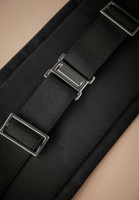 Correa negra ajustable con hebillas de metal plateado sobre fondo de tela negra texturizada.