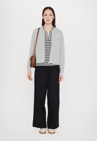 Šedá bomber bunda s přední kapsou, pruhované tričko pod ní, volné černé kalhoty a béžové boty. Součástí je hnědá crossbody taška.