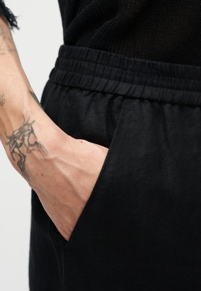 Μαύρο λινό παντελόνι με ελαστική μέση και πλαϊνή τσέπη. Στενή λήψη δείχνει ένα χέρι με ορατό τατουάζ να ξεκουράζεται στην τσέπη.