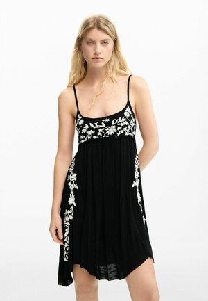 Fiatal nő fekete, ujjatlan ruhában, fehér virágmintás hímzéssel, egyszínű fehér háttér előtt áll.