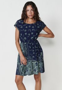 Rochie bleumarin cu imprimeu paisley verde, cu mâneci scurte și talie elastică. Fustă evazată cu un design stratificat și detalii cu tiv curbat.