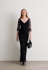 Robe en dentelle noire avec des manches trois-quarts, design ajusté et décolleté en V, accompagnée d'un petit sac à main noir texturé.