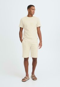Ensemble chemise et short beige texturé, avec manches courtes et encolure ronde, associé à des sandales marron clair sur un fond uni.