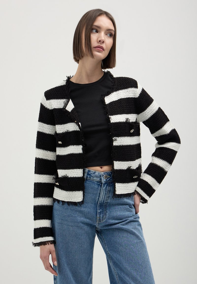 Cardigan a righe bianco e nero in maglia con bordi sfrangiati, con un design accorciato e bottoni, indossato sopra una maglietta nera e jeans a vita alta.