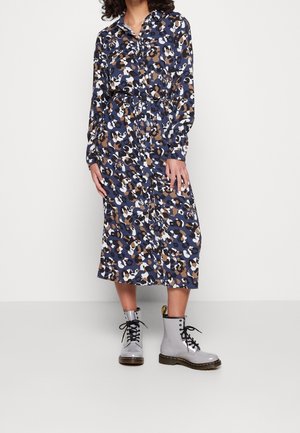 Vrouw die een midi-jurkje met abstract patroon in blauw, bruin, wit en zwart draagt, met een tailleband en lichtgrijze veterschoenen tot de enkel.