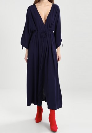 Vrouw die een lange marineblauwe wikkeljurk draagt met wijde mouwen en rode enkellaarzen, staand tegen een effen lichtgrijze achtergrond.