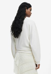 Biely sweatshirt s nižšími ramennými švami, jemnou textúrou a rebrovanými manžetami, kombinovaný s bielou pletenou sukňou so vzorovými detailmi a strapcami.