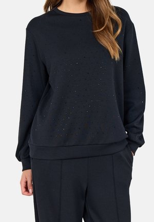 Kobieta ubrana w czarną, długorękawową bluzę z małymi błyszczącymi ozdobami oraz dopasowane czarne spodnie, ręce w kieszeniach.