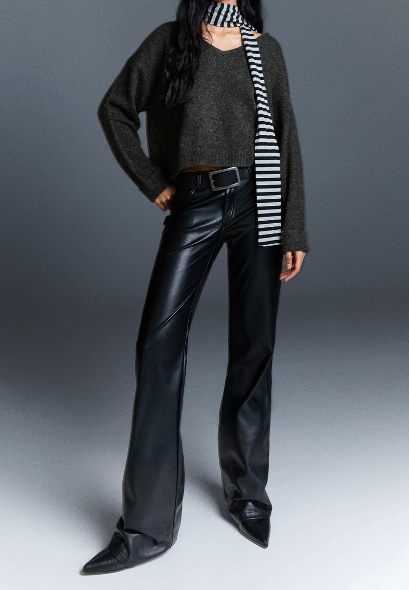 Femme portant un pantalon en cuir noir ample, un pull court gris foncé, une écharpe rayée noire et blanche, et des chaussures pointues noires sur fond gris.