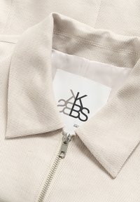 Beige zip-up jacka med en strukturerad krage, ribbad textur och en etikett med en logotyp. Silverdragkedja fram.