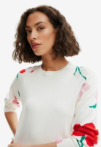 Witte trui met bloempatronen in tinten van roze, rood en groen op de mouwen, met een ronde hals en geribde manchetten.