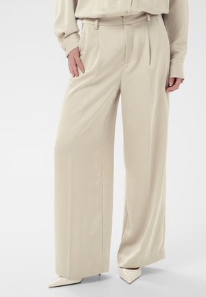 Béžové široké nohavice s vysokým pásom nosené s rovnakou blúzkou a lodičkami so špicatou špičkou, ruky odpočívajúce pri vreckách.