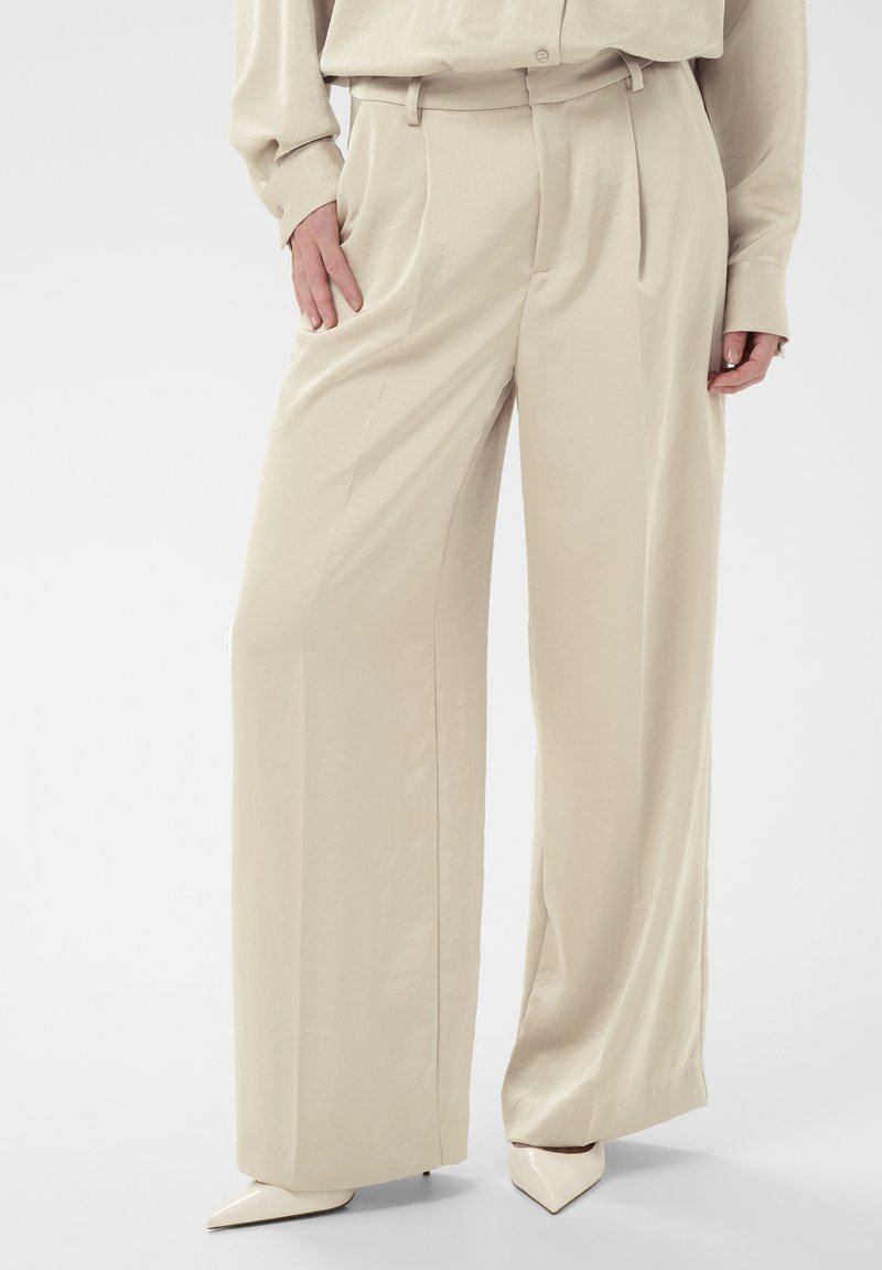 Pantalon beige taille haute à jambes larges porté avec un chemisier assorti et des escarpins à bout pointu, les mains reposant près des poches.