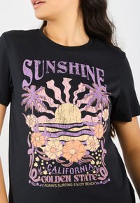 Svart T-skjorte med et fargerikt grafikk av en sol, bølger og blomster med teksten: "Sunshine California Golden State" i lilla og ferskenfarger.