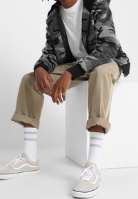 Sudadera con capucha de camuflaje gris, camiseta blanca, pantalones beige con puños, calcetines blancos con rayas grises y zapatillas gris claro con cordones blancos.