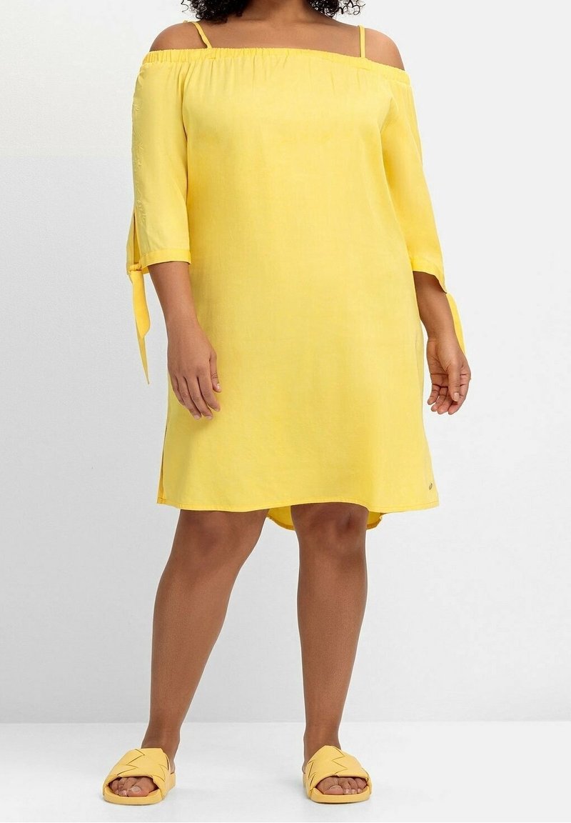 Femme portant une robe jaune épaules dénudées avec des manches trois-quarts et des sandales jaunes assorties, debout sur un sol blanc.