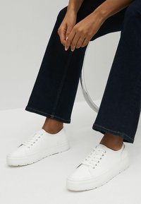 Adidași albi cu șireturi, cu un finisaj neted, vârf rotunjit și o talpă groasă. Asortați cu blugi cu picior larg de culoare albastru închis.