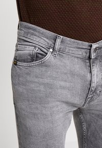 Primer plano de una persona vistiendo jeans de mezclilla gris desgastados y una camisa de color marrón oscuro texturizada por dentro.
