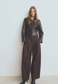 Brązowa skórzana krótka kurtka z czarnymi guzikami, zestawiona z brązowymi spodniami z wysokim stanem i szerokimi nogawkami. Modelka ma na sobie czarne botki na obcasie.