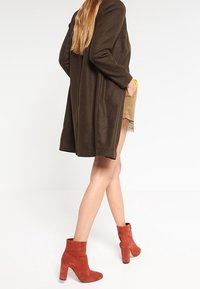 Manteau long en mélange de laine marron, avec une coupe décontractée, associé à un short beige et des bottes à talons épais en daim rouge.