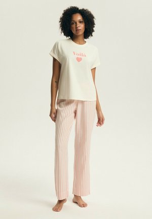 Kobieta o kręconych włosach, ubrana w białą koszulkę z napisem „Voilà” i sercem, w połączeniu z białymi spodniami w pionowe różowe paski, stojąca boso.