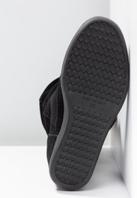 Botte en daim noir avec une semelle en caoutchouc texturée présentant un motif de bande de roulement. Le nom de la marque et la taille sont embossés sur la semelle.