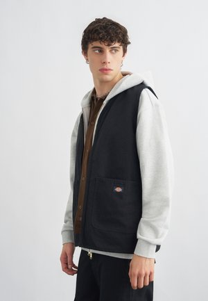 Młody mężczyzna w szarej bluzie z kapturem i czarnej kamizelce z przednimi kieszeniami, patrzący na bok na tle jednolitej białej ściany.