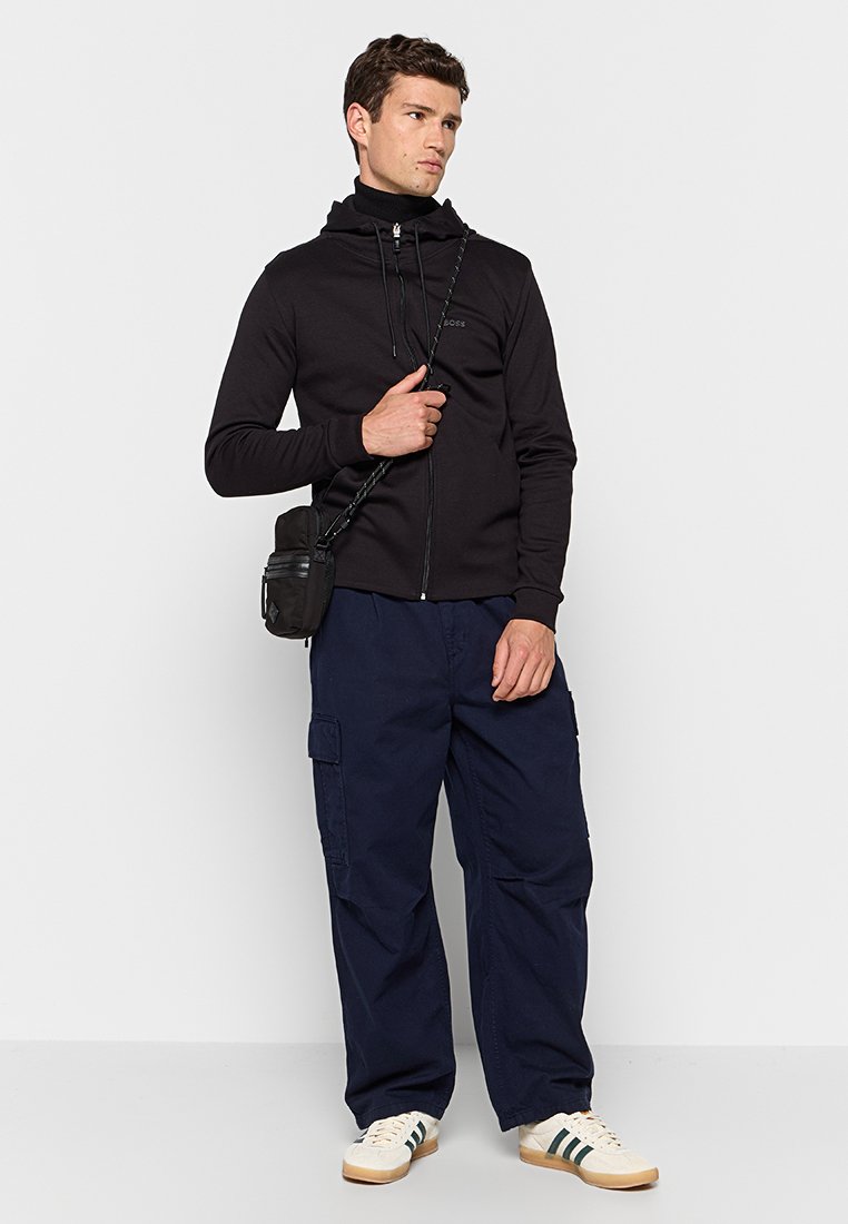 Zwarte zip-up hoodie met hoge kraag, marineblauwe cargo broek en lichte sneakers met groene strepen. Model houdt een kleine zwarte tas vast.