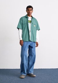 Grön t-shirt med kort ärm över en vit skjorta med lång ärm, parat med lösa blå denimjeans och grå sneakers. Enkelt design med fickor.