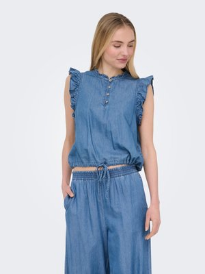 Femme portant un haut en jean bleu sans manches avec des volants aux épaules et un pantalon large taille haute assorti, debout avec une main dans la poche.