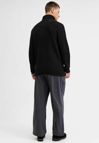 Zwarte gebreide oversized trui met een hoge kraag, textuurpatroon, geribbelde zoom, gecombineerd met losse grijze broeken en zwarte schoenen.
