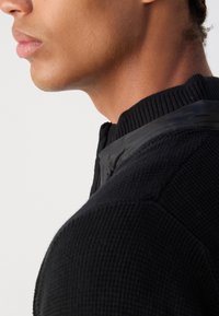 Black textured sweater with a ribbed collar, featuring a contrasting smooth black accent at the neckline. Close-up view of the shoulder and neck area.