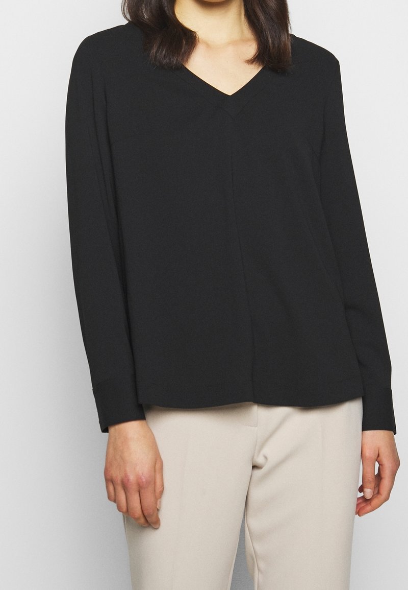 Femme portant un haut noir à manches longues avec un col en V et un pantalon beige, vue des épaules aux hanches sur un fond uni.