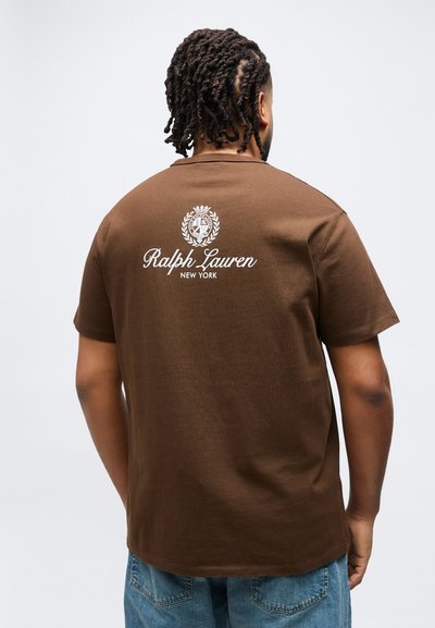 Άνδρας με πλεξούδες, φοράει καφέ μπλουζάκι Ralph Lauren με λευκό κείμενο λογότυπου στο πάνω μέρος της πλάτης και μπλε τζιν παντελόνι.