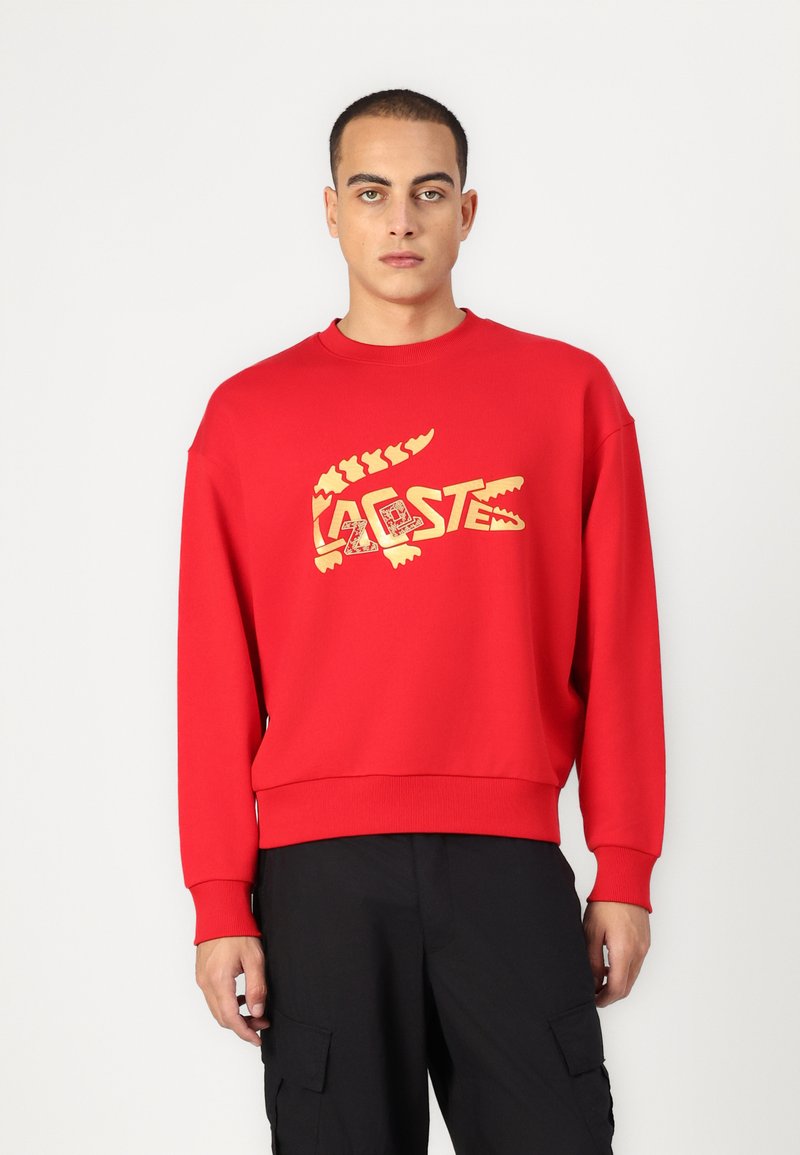 Felpa rossa in cotone, con un grande logo di un coccodrillo dorato e testo sul davanti. Maniche e orlo a coste con una vestibilità comoda.