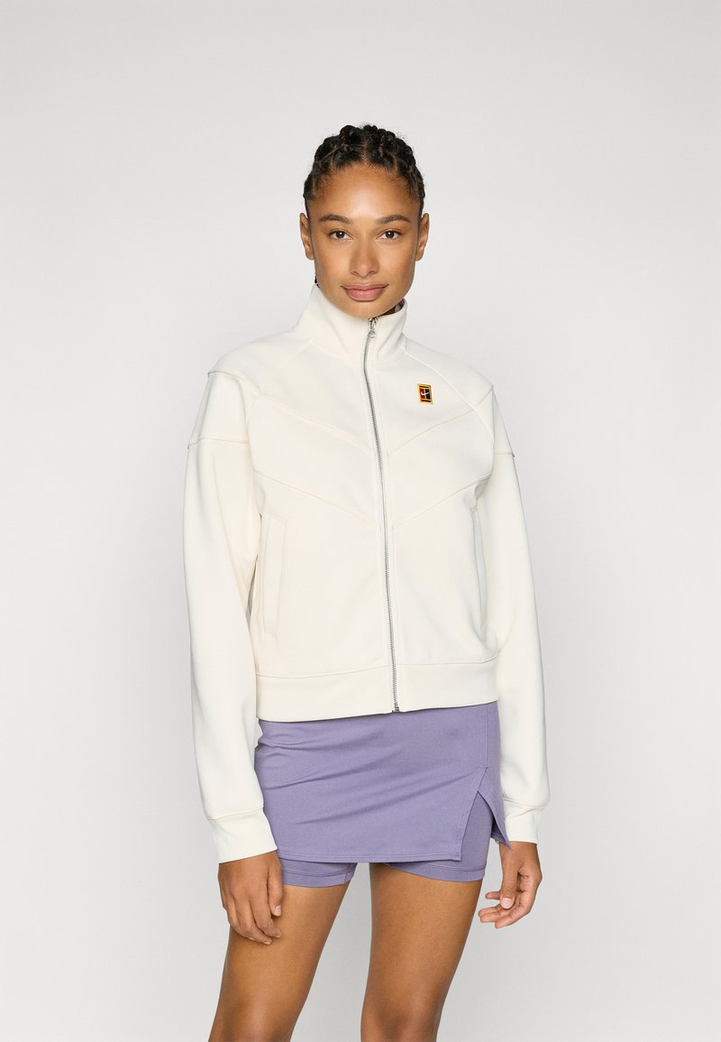 Lyscreme zip-up jakke med høj krave, der har chevron-sømme. Båret over en lavendel nederdel, der fremhæver en blød tekstur og et enkelt design.