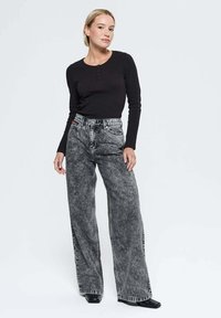 Femme debout portant un haut à manches longues noir, un jean large à taille haute délavé avec effet acide, et des chaussures noires à bout carré, sur fond blanc.