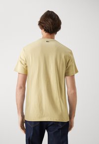Camiseta de algodón de manga corta, color amarillo claro, con cuello redondo y un pequeño logo verde en la parte trasera. Textura suave y lisa.