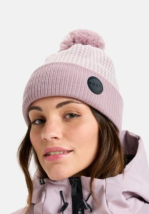 Frau trägt eine rosa gestrickte Mütze mit Bommel und eine lavendelfarbene Jacke, schaut nach vorne und lächelt leicht.