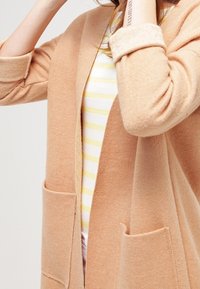 Hellbeige Cardigan mit lässiger Passform, Material aus Wollmischung, offenes Vorderteil, zwei Seitentaschen und aufgerollte Manschetten über einem gestreiften gelb-weißen Oberteil.
