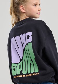 Sweatshirt noir avec un grand graphique coloré au dos, présentant "Nyc" en violet et "Sport" en vert, ainsi qu'un texte orange en dessous.