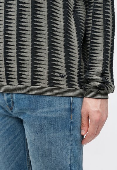 Πουλόβερ με ρίγες σε μαύρο και γκρι χρώμα με υφασμένους σχεδιασμούς, φορεμένο με τζιν παντελόνι ανοιχτού μπλε χρώματος. Διαθέτει μανσέτες και στρίφωμα με ριμπ ύφανση.