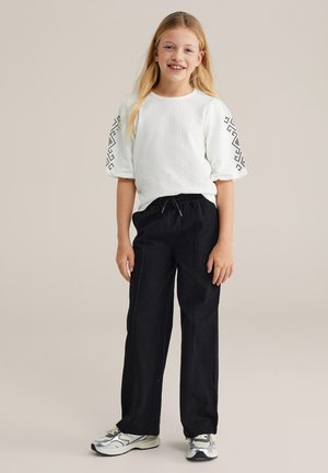 Bela kratka majica z reliefno teksturo in črnim geometrijskim vzorcem na rokavih, kombinirana z ohlapnimi črnimi hlačami in sivimi supergami.