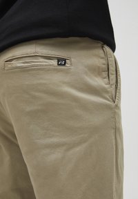 Beige bomullbyxor med en mjuk textur, som har en liten svart logotyp på bakfickan och sidofickor.