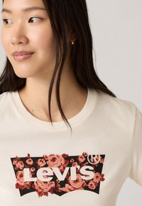 T-shirt couleur crème arborant un graphique floral dans des teintes de rose et de rouge, avec un fond noir et le logo texte "Levi's" en blanc.
